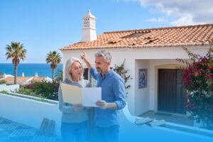 Älteres Paar steht mit Dokumentenmappe vor seinem weißen Haus an der Algarve und begutachtet die vom Sturm beschädigten Dachziegel