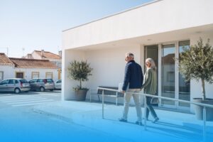 Ein Paar um 65 geht über eine Rampe zum gläsernen Eingang eines modernen weißen Gesundheitszentrums in einer portugiesischen Kleinstadt